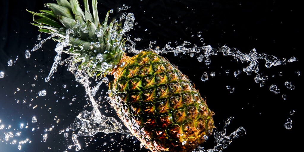 Eine frische Ananas fliegt durch die Luft, umgeben von fließendem Wasser, auf einem schwarzen Hintergrund. Die Wasserspritzer reflektieren Licht und erzeugen ein dynamisches Bild, das die Frische und Saftigkeit der Ananas betont. Ideal für einzigartige Wandbilder.