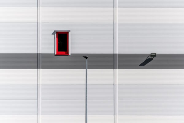 Roland Seichter Fotografie - Einzigartige Wandbilder - Minimalistische Fotografie - Eine grau-weiße Fassade mit einem rot gerahmten Fenster