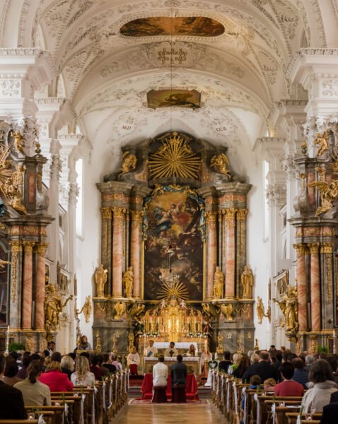 Roland Seichter Fotografie - Hochzeitsfotografie - Feiereliches Hochzeitsmotiv in einer barocken Kirche, die Gäste sitzen links uns rechts in den Sitzreihen, vorne sitzen Braut und Bräutigam, die von hinten fotografiert sind