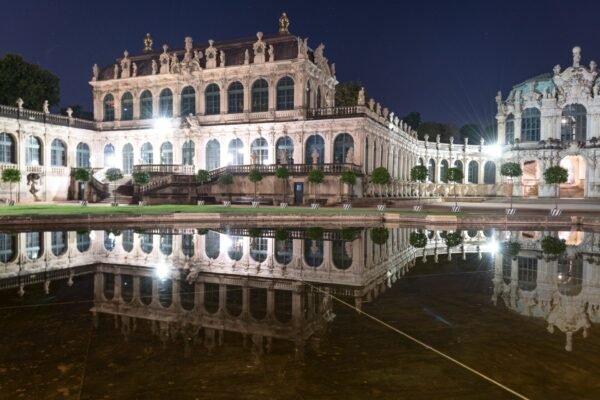 Historisches Barockgebäude bei Nacht, beleuchtet mit Wasserspiegelung und Statuen entlang Fassade.