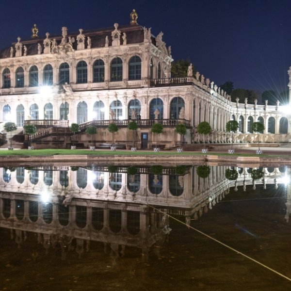 Historisches Barockgebäude bei Nacht, beleuchtet mit Wasserspiegelung und Statuen entlang Fassade.