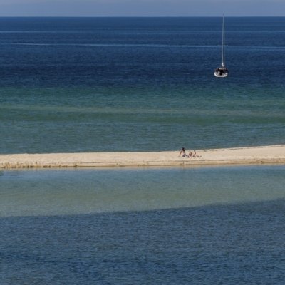 Segelboot auf Meer, zwei Personen auf Sandbank, blaues Wasser, ruhige und sonnige Szene.