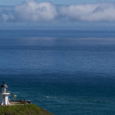 Leuchtturm auf grünem Hügel am Meer mit bewölktem Himmel und Personen in der Nähe.