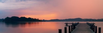 Roland Seichter Fotografie - Landschaftsfotografie - Morgens am Wörthsee über eine Seebrücke aufs Wasser fotografiert. Die morgendliche Sonne erzeugt ein rotes Licht