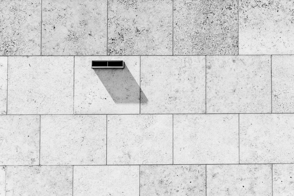Roland Seichter Fotografie - Minimalistische Fotografie - Eine Steinfassade mit einem Belüftungsausgang, großer Schattenwurf