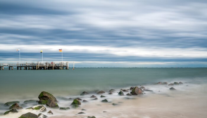 Roland Seichter Fotografie - Einzigartige Wandbilder - Fotografie mit Wasser - Ein Schiffststeg von links ins Wasser hinenragend, Steine im Vordergrund. Durch Langzeitbelichtung verschwimmen Wellen und Wolken