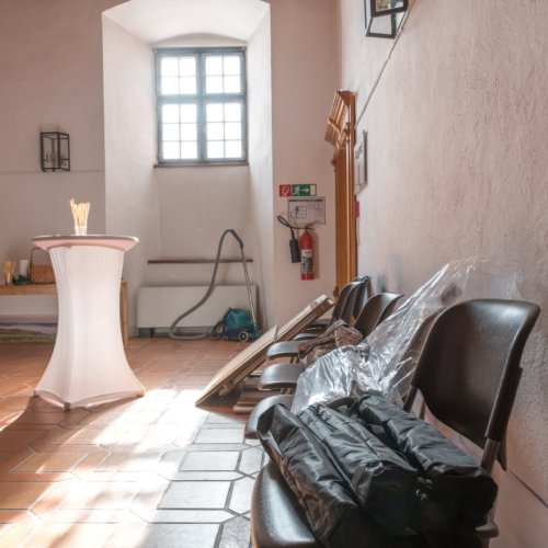 Raum mit Fliesenboden, Tisch, Stühlen; Fenster, Bilder, Feuerlöscher, Person in der Ecke.