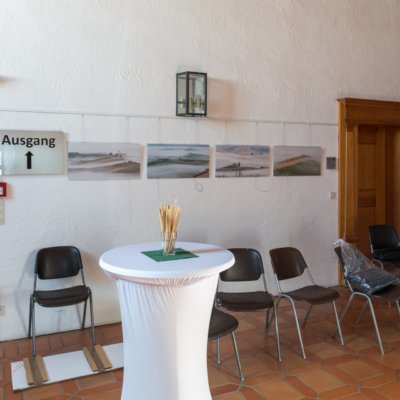 Raum mit weißen Wänden, Fotos, "Ausgang"-Schild, Klappstühlen, Stehtischen und Terrakotta-Boden.