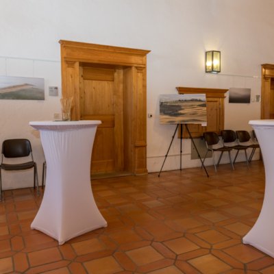 Ausstellungsraum mit weißen Stehtischen, Stühlen, Landschaftsfotos an Wand und Holzdetails.