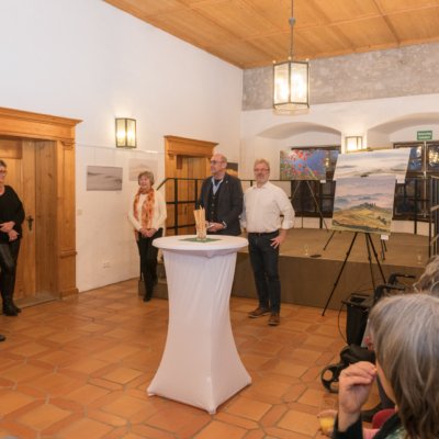 Menschen im Ausstellungsraum mit Gemälden auf Staffeleien und warmer Beleuchtung.