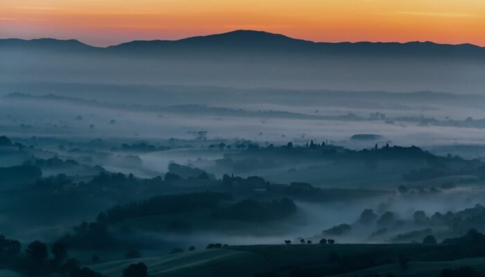 Roland Seichter Fotorgafie - Landschaftsfotografie - Morgenstimmung einer toskanischen Landschaft mit orange gefärbtem Himmel und einem dunklem Tal, in dem sich Nebel verfangen hat