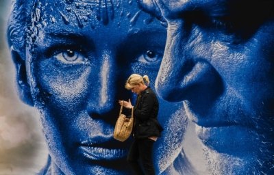 Roland Seichter Fotografie - Street Fotografie -Eine Dame steht vor einem großen Filmplakat, auf dem blauer Gesichter durchdringend schauen. Die Dame schaut in ihrer Handtasche etwas nach. Es sieht aus, als ob die Personen des Plakates in die Tasche schauen