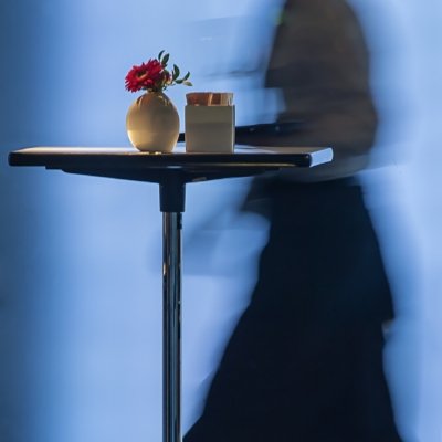 Roland Seichter Fotografie - Fotowettbewerbe - Ein Stehtisch mit eine kleinen Blumenvase darauf. Im Hintergrund läuft eine Servivekraft von rechts nach links. Durch Langzeitbelichtung ist diese Servicekraft verschwommen dargestellt