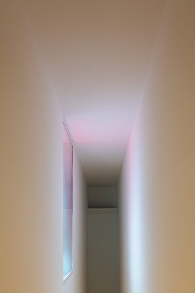 Abstrakte Architekturfotografie eines schmalen, weißen Flurs mit weichem, violett-rosa Lichtschein an der Decke