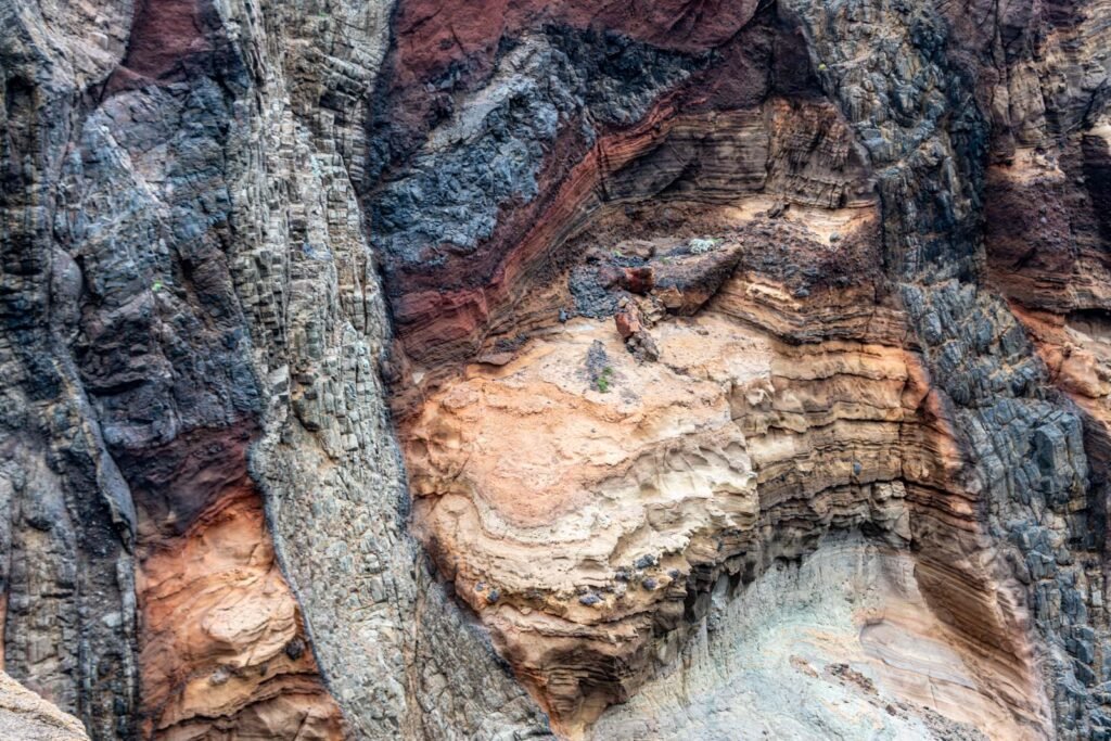 Detailaufnahme einer zerklüfteten Felswand mit deutlich erkennbaren geologischen Schichten in Rotbraun, Ocker und Graublau