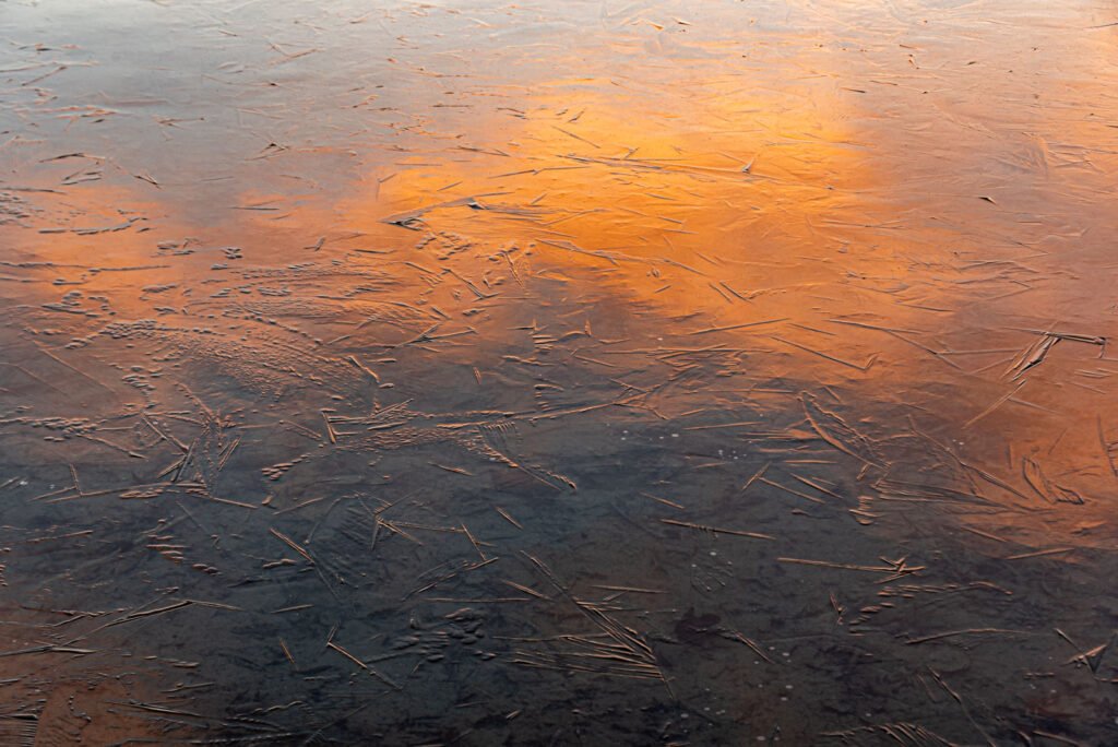 Abstrakte Fotokunst von feinen Eisstrukturen auf einer Wasseroberfläche mit orangefarbenen Lichtreflexionen
