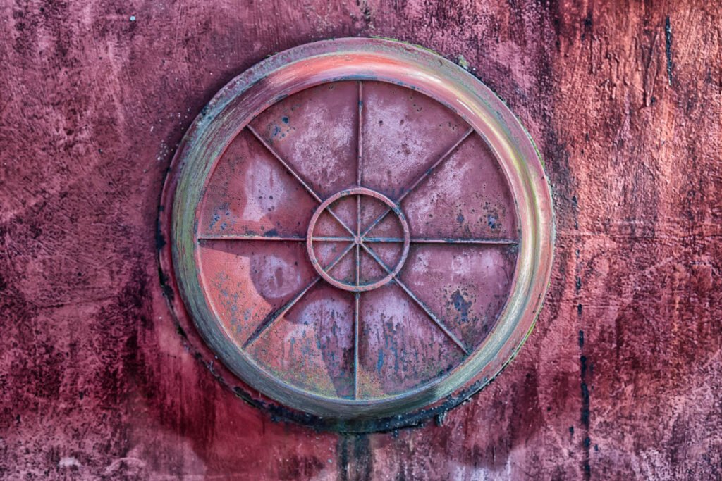 Abstrakte Nahaufnahme eines runden, symmetrisch unterteilten Metallelements mit starker roter Patina und verwitterter Oberflächenstruktur vor einer farblich passenden Wand
