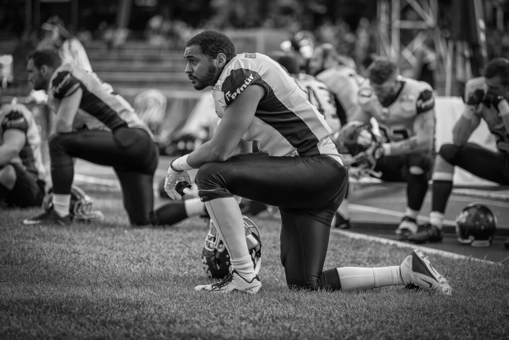 Emotionale schwarz-weiß Aufnahme eines Football-Spielers, der erschöpft und enttäuscht am Spielfeldrand kniet.