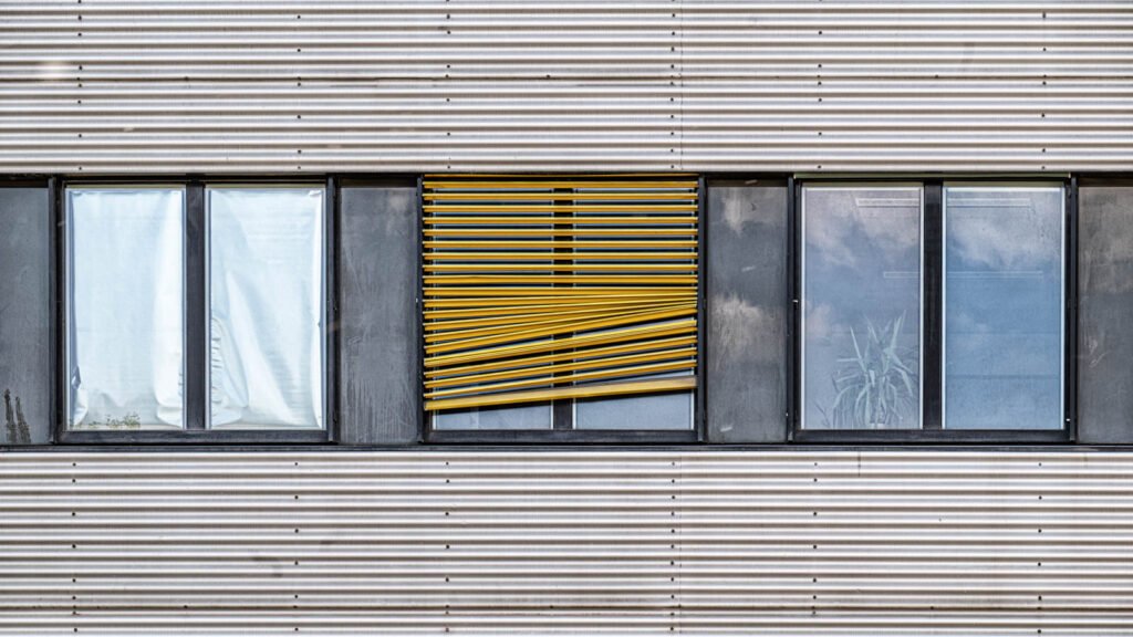 Detailaufnahme einer Wellblechfassade mit einer auffälligen, defekten gelben Jalousie im Fenster.