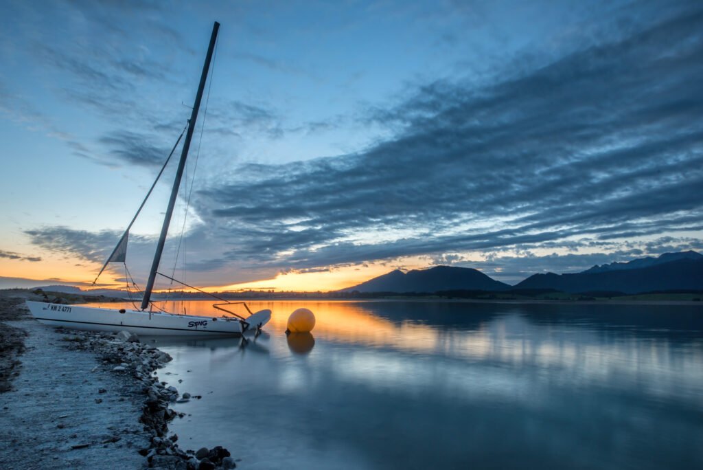 Morgenstimmung am Forggensee: Segelboot am Ufer beim Übergang von der Blauen zur Goldenen Stunde.