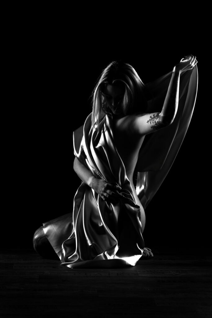 Künstlerische Schwarz-Weiß-Aktfotografie einer Frau mit einem fließenden hellen Tuch und starken Kontrasten.