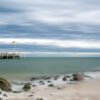 Das Wandbild Auf Föhr zeigt eine ruhige Strandlandschaft mit Buhnen im Vordergrund und einer Seebrücke unter einem weiten, dramatischen Wolkenhimmel