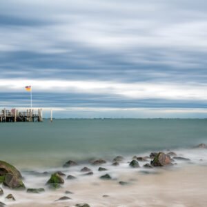 Das Wandbild Auf Föhr zeigt eine ruhige Strandlandschaft mit Buhnen im Vordergrund und einer Seebrücke unter einem weiten, dramatischen Wolkenhimmel