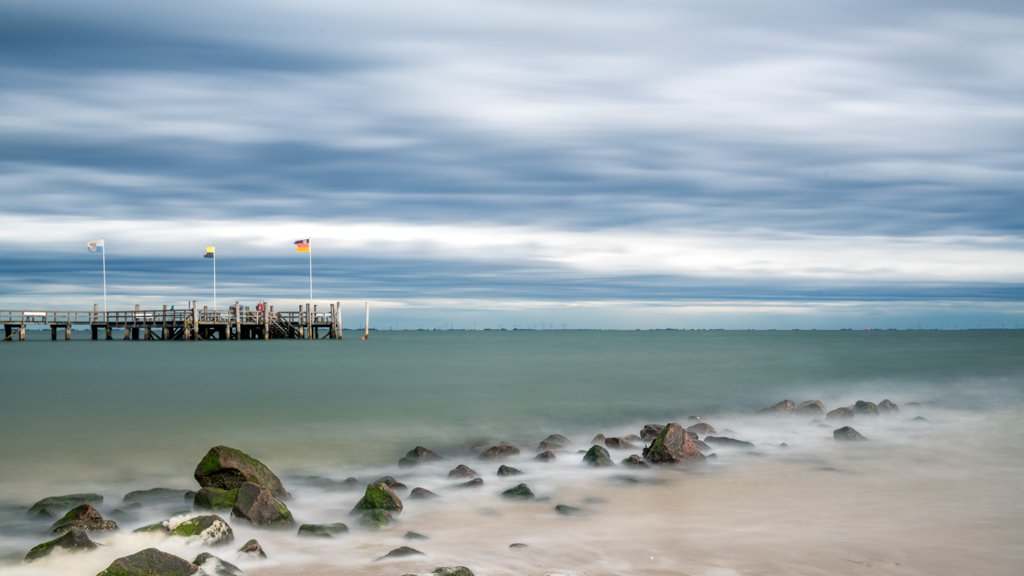 Das Wandbild Auf Föhr zeigt eine ruhige Strandlandschaft mit Buhnen im Vordergrund und einer Seebrücke unter einem weiten, dramatischen Wolkenhimmel