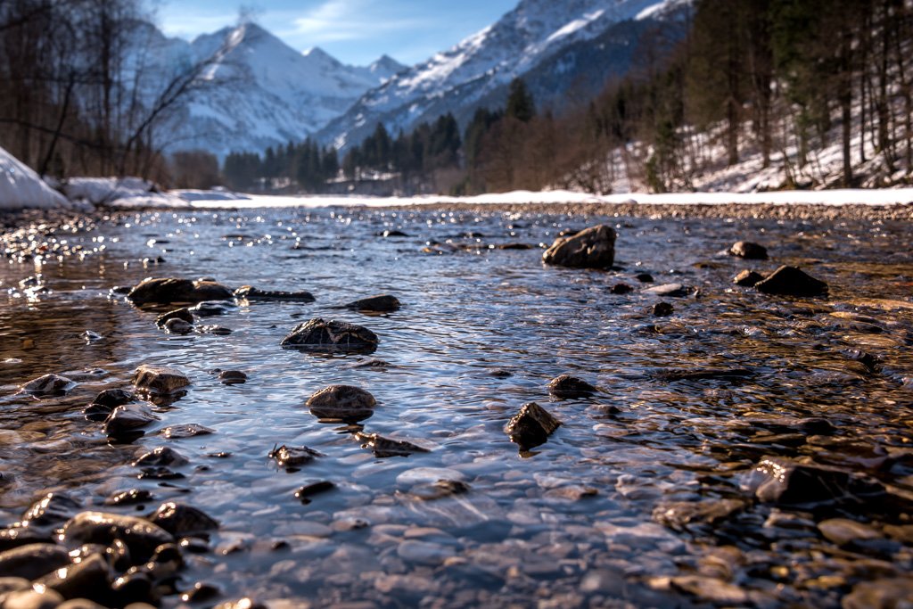 Das Wandbild Bergfluss zeigt klares, glitzerndes Wasser eines Gebirgsbachs im Vordergrund und schneebedeckte Gipfel im Hintergrund