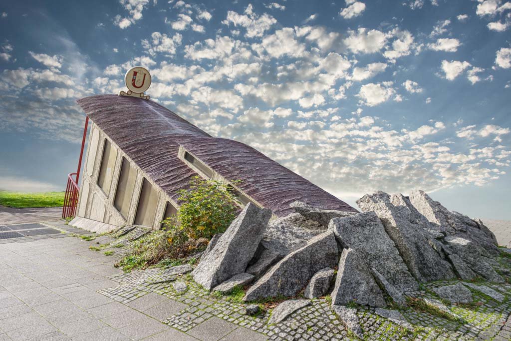Das Composing Downstairs Underground zeigt einen surrealen U-Bahn-Aufgang, der organisch aus einem Felsen unter einem weiten Wolkenhimmel ragt.