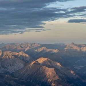 Fotokunst Die Gipfel werden wach Zugspitze: Panorama beleuchteter Berggipfel vor graublauem Wolkenhimmel bei Sonnenaufgang.