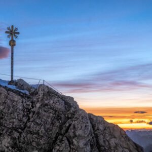 Fotokunst Gipfelkreuz Zugspitze: Das Wahrzeichen auf dem Gipfelfelsen im Schatten vor einem orange-gelben Morgenhimmel.