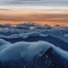 Fotokunst Morgendämmerung Zugspitze: Dunkle Bergsilhouetten über denen sich eine massive, weiße Wolkenwelle vor orangefarbenem Himmel bricht.