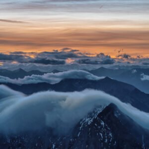 Fotokunst Morgendämmerung Zugspitze: Dunkle Bergsilhouetten über denen sich eine massive, weiße Wolkenwelle vor orangefarbenem Himmel bricht.