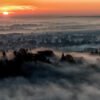 Das Wandbild Mystischer Nebel zeigt eine Stadt im Tal, die bei Sonnenaufgang fast vollständig in dichten Bodennebel gehüllt ist, fotografiert aus einer erhöhten Perspektive