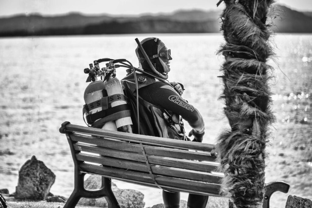 Schwarzweiß-Fotografie eines Tauchers in voller Ausrüstung auf einer Parkbank am See.