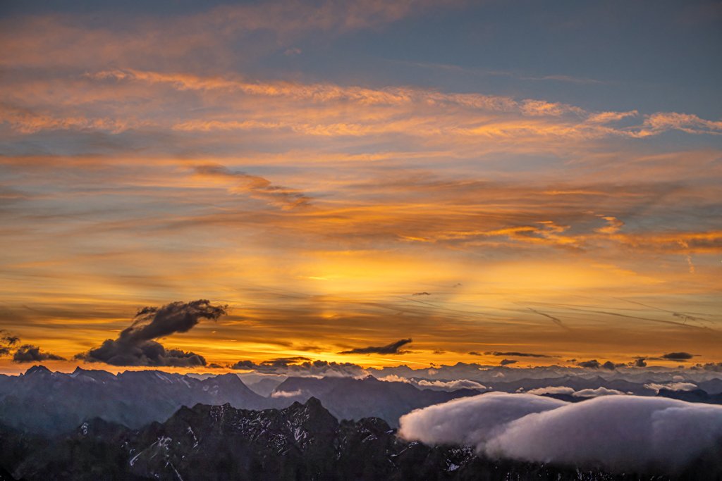 Fotokunst Von der Zugspitze: Dramatischer Sonnenaufgang mit intensiv orange-gelb leuchtenden Wolken über dunklen Bergsilhouetten.