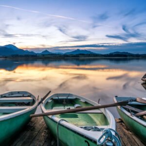 Das Wandbild Abend am Hopfensee zeigt mehrere Ruderboote im Vordergrund an einem Holzsteg, deren Spiegelung sich zusammen mit den fernen Alpen im ruhigen Wasser bei Sonnenuntergang bricht.