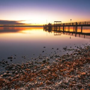 Das Wandbild Ammersee abends zeigt einen langen Holzsteg, der in den spiegelglatten See führt, während der Horizont in warmen Gold- und Orangetönen leuchtet und das Ufer mit herbstlichem Laub bedeckt ist.