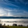 Das Wandbild Blick vom Auerberg am Morgen zeigt eine weite Sicht über ein dichtes Nebelmeer im Tal auf die Allgäuer Alpenkette unter einem dynamischen, blau-weißen Wolkenhimmel.