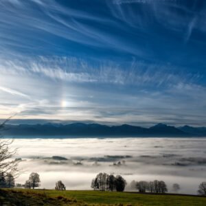 Das Wandbild Blick vom Auerberg am Morgen zeigt eine weite Sicht über ein dichtes Nebelmeer im Tal auf die Allgäuer Alpenkette unter einem dynamischen, blau-weißen Wolkenhimmel.