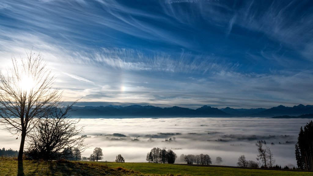 Das Wandbild Blick vom Auerberg am Morgen zeigt eine weite Sicht über ein dichtes Nebelmeer im Tal auf die Allgäuer Alpenkette unter einem dynamischen, blau-weißen Wolkenhimmel.