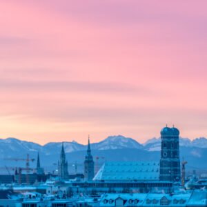 Das Panorama-Wandbild Frauenkirche am Morgen zeigt die Münchner Skyline mit Alpenpanorama unter einem rosa leuchtenden Winterhimmel.
