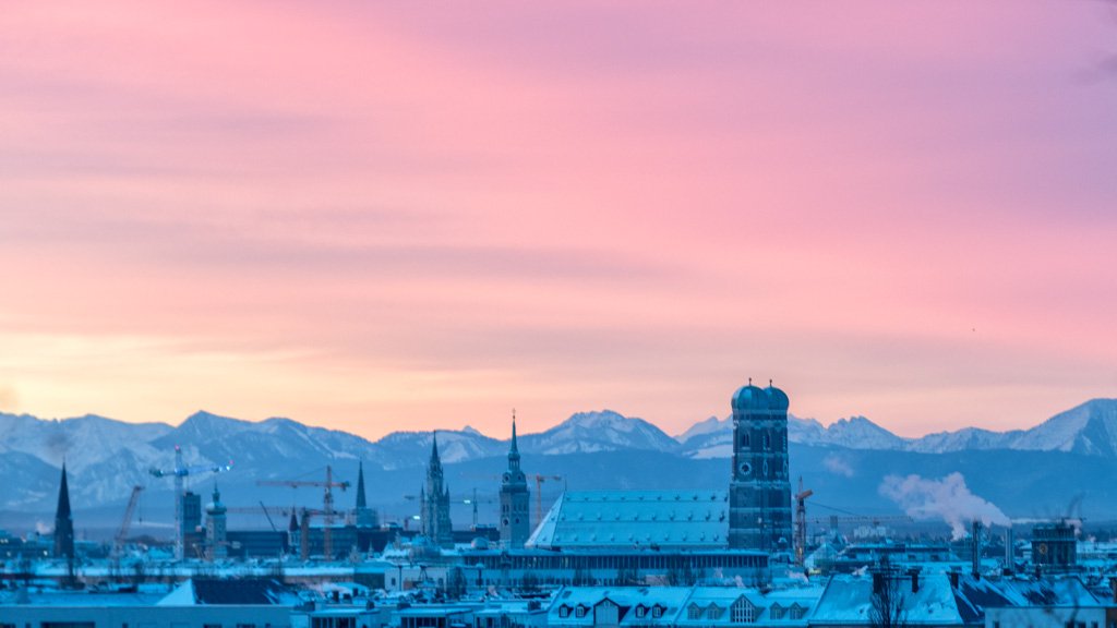 Das Panorama-Wandbild Frauenkirche am Morgen zeigt die Münchner Skyline mit Alpenpanorama unter einem rosa leuchtenden Winterhimmel.
