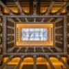 Das Wandbild Im Innenhof zeigt eine symmetrische Architekturaufnahme eines historischen Innenhofs mit Blick nach oben zu einem Glasdach.