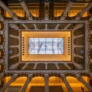Das Wandbild Im Innenhof zeigt eine symmetrische Architekturaufnahme eines historischen Innenhofs mit Blick nach oben zu einem Glasdach.