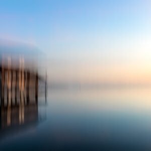 Das Wandbild Lake House zeigt eine abstrakte, durch Kamerabewegung verschwommene Aufnahme eines Bootshauses auf einem ruhigen See in sanften Blau- und Goldtönen.