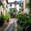 Das Wandbild Mediterraner Innenhof zeigt eine malerische italienische Gasse mit Kopfsteinpflaster, üppigen Grünpflanzen und historischen Fassaden im 3:2 Format.