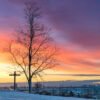 Das Wandbild Morgens vom Olympiaberg zeigt einen verschneiten Hügel mit markantem Baum und Gipfelkreuz vor einem farbenprächtigen Sonnenaufgang über München.