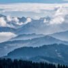 Das Wandbild Mountain Mist zeigt eine tief gestaffelte Berglandschaft im Allgäu, bei der dunkle Nadelwälder im Vordergrund in blau-graue Bergsilhouetten übergehen, die von weißen Wolken und Nebel umspielt werden.